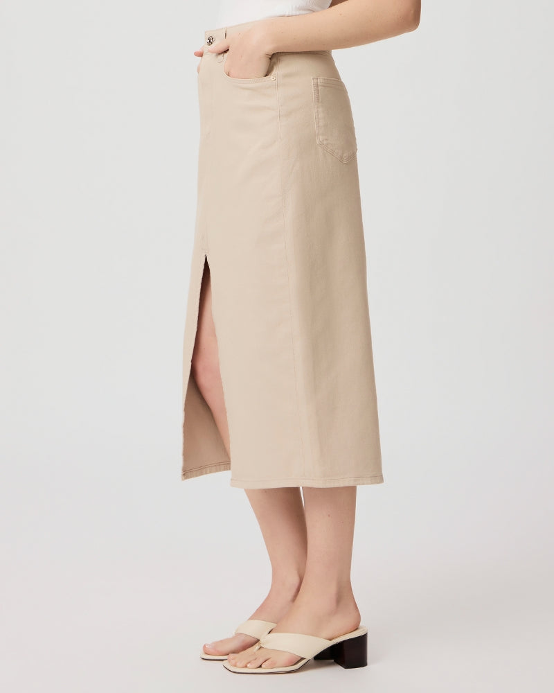 A person stands wearing a Paige Angela midi skirt in Soft Beige with a high waist side slit, paired with white sandals, against a light background.