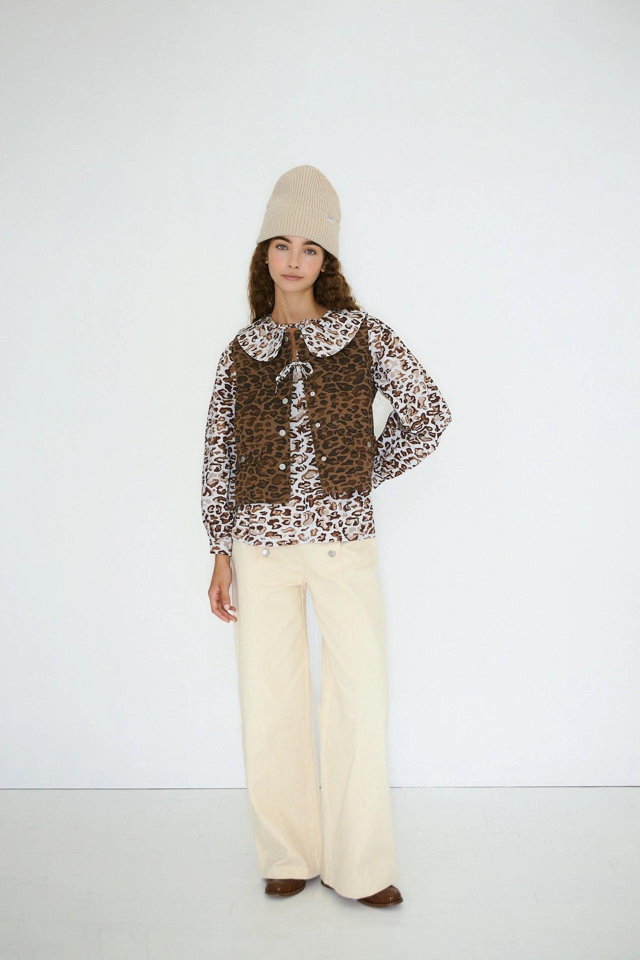 A person stands against a white wall wearing the Tiny Big Sister Animal Print Vest - Mocha over a leopard print blouse with a large collar, beige knit hat, cream wide-leg pants, and brown shoes.