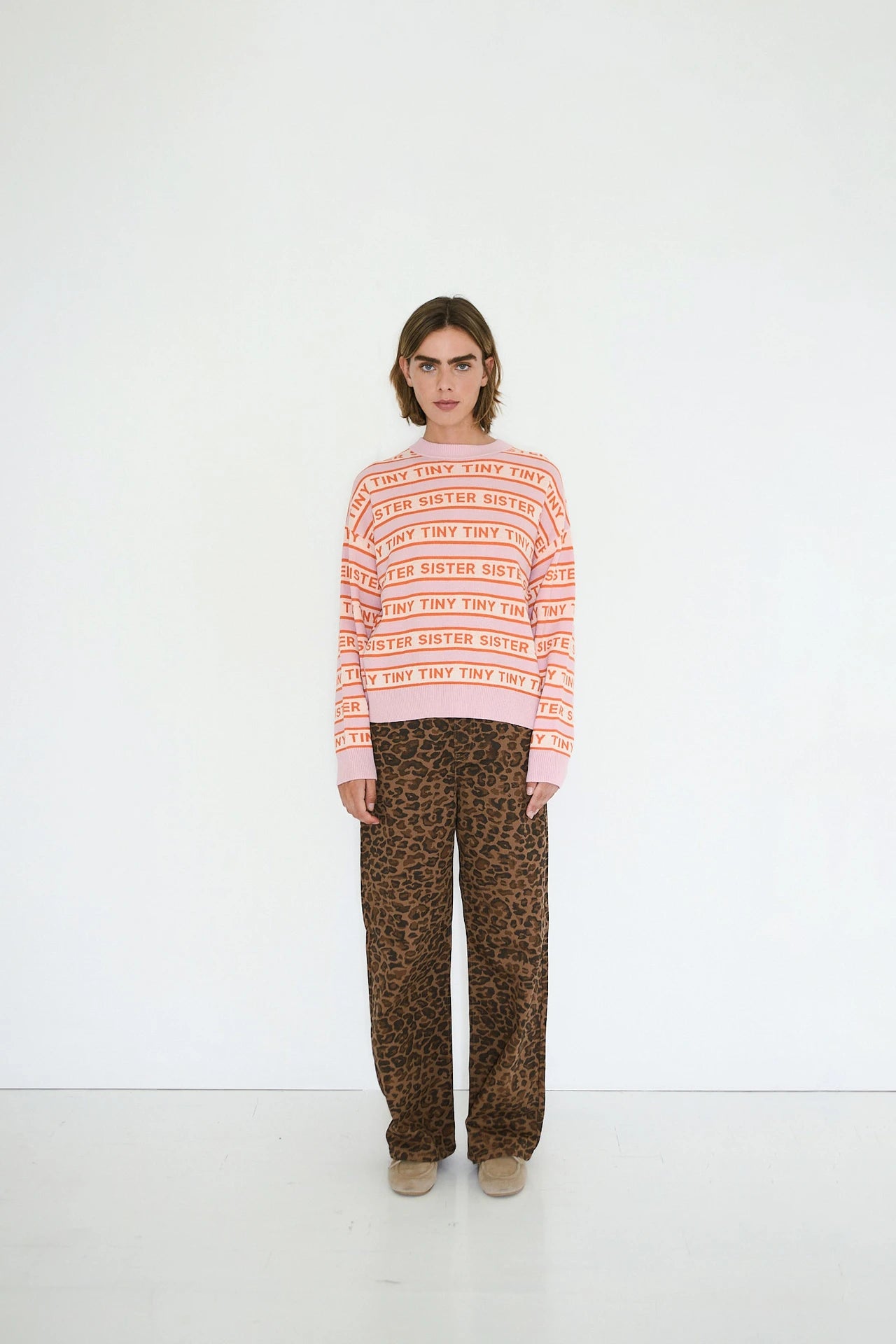 A person stands against a plain white wall, wearing pink and red striped sweater text, paired with Tiny Big Sister’s Berta Animal Print Jeans in Mocha.