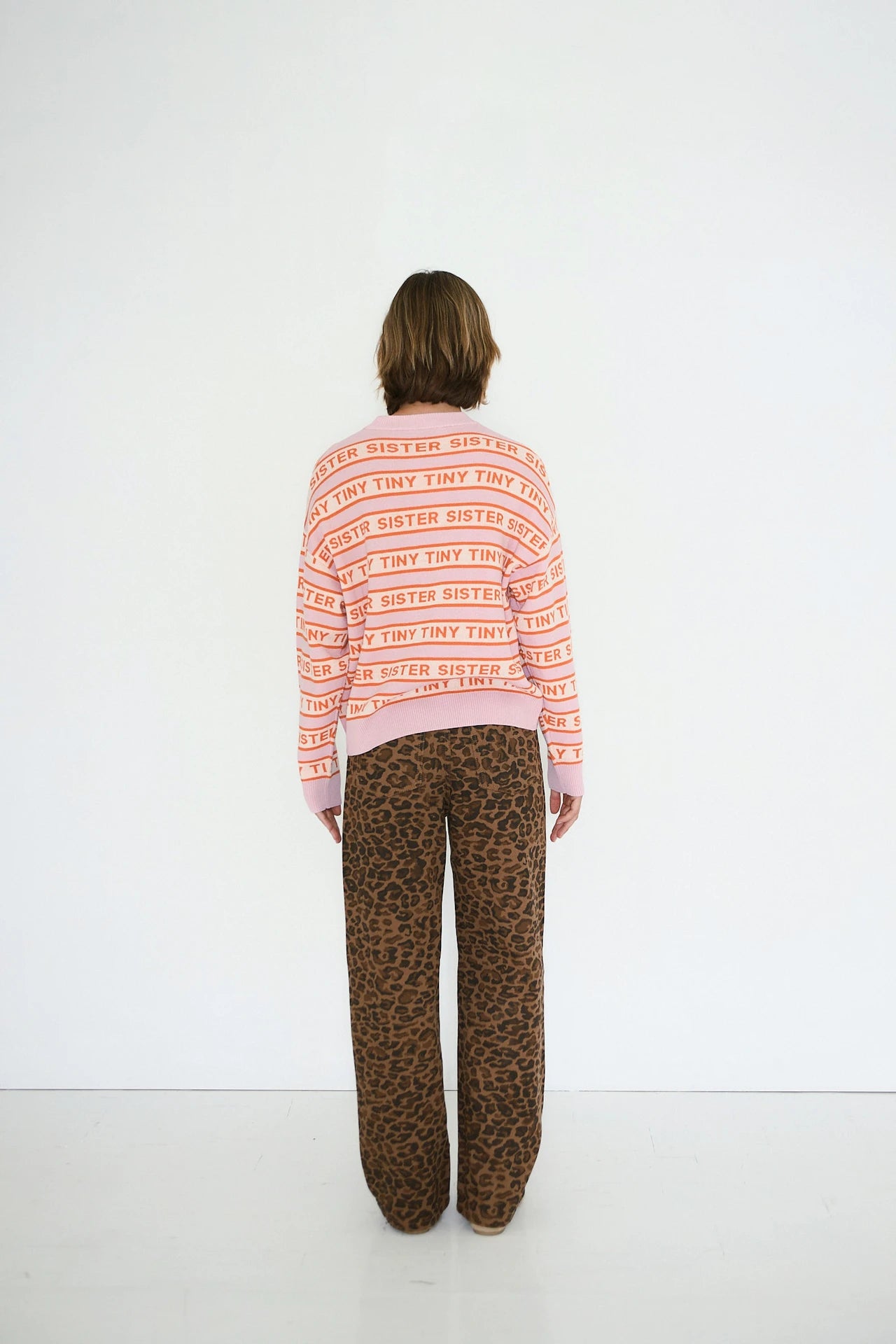 A person stands facing a white wall, wearing a pink sweater with repeated "TINY SISTER" text and Tiny Big Sister's Berta Animal Print Jeans in mocha.