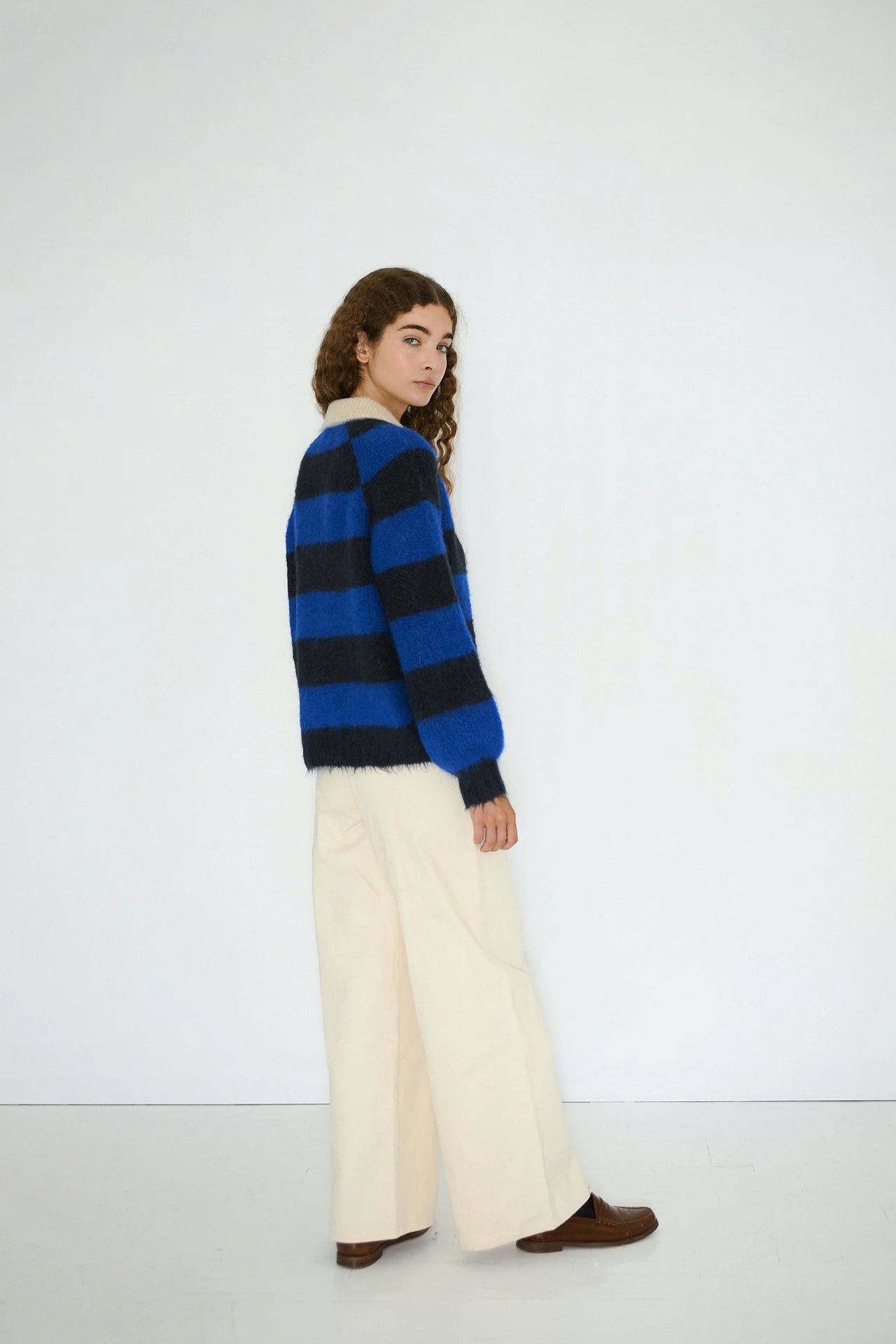 A person with long curly hair wears Tiny Big Sister's Frank Striped Polo Jumper in navy, paired with wide-leg cream pants and brown shoes, standing against a plain white background.