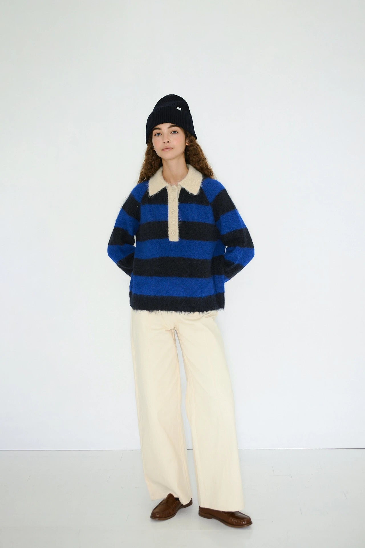 A person stands against a plain white wall wearing the Tiny Big Sister Frank Striped Polo Jumper in navy, paired with cream wide-leg pants, brown shoes, and a navy beanie.