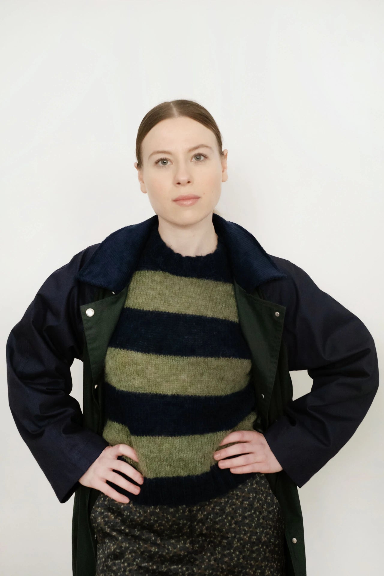 A light-skinned person with brown hair stands hands on hips against a white background, wearing a dark jacket and the Tiny Big Sister Oversized Striped Sweater in Navy/Khaki, made with sustainable manufacturing.