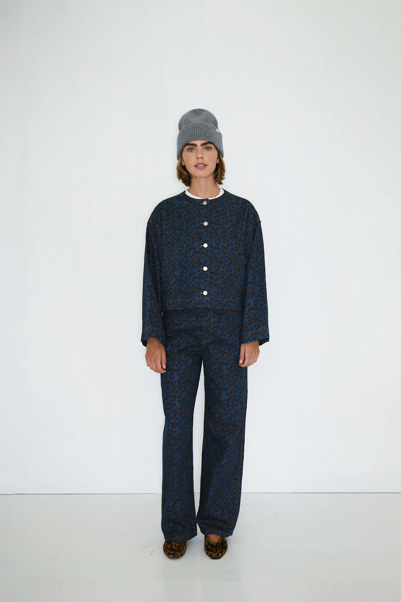 A person stands against a plain white wall wearing the Tiny Big Sister Flowers Cropped Jacket in Navy, paired with matching pants, a grey beanie, and leopard print shoes.