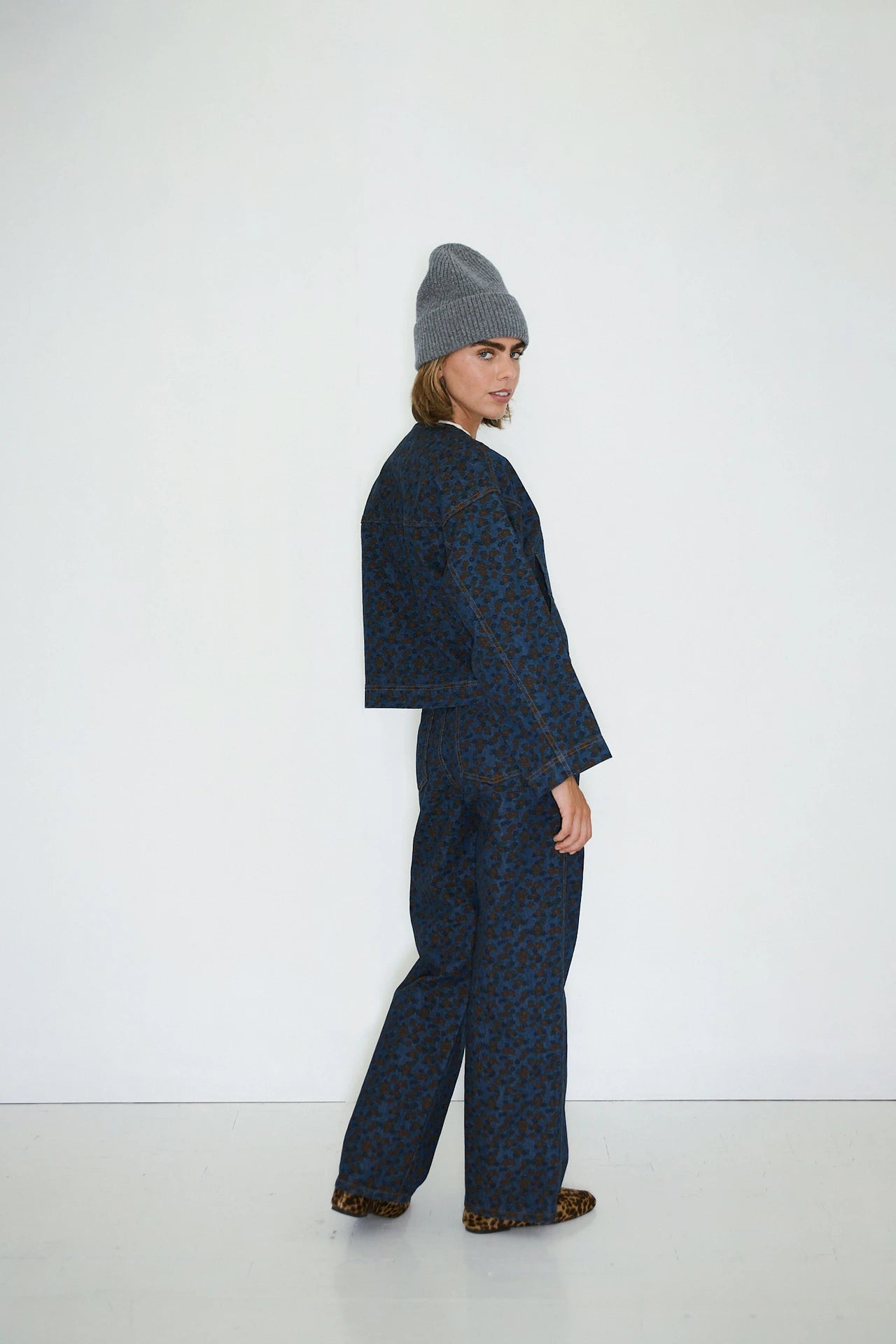 Person standing sideways against a white wall, wearing a grey knit beanie, the Tiny Big Sister Flowers Cropped Jacket in Navy, and leopard print shoes.
