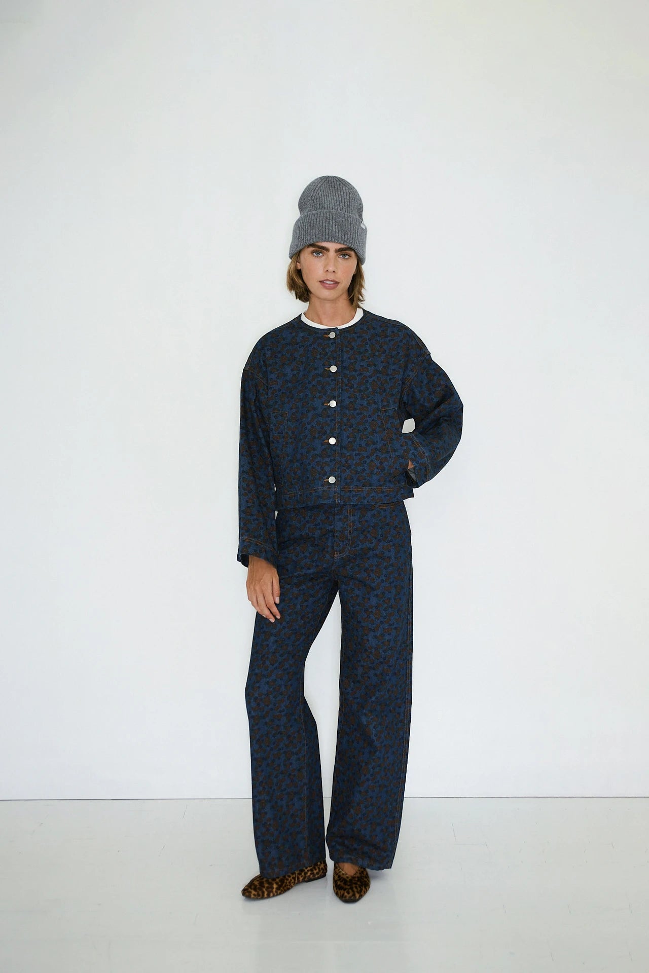 A woman stands against a plain white wall wearing Tiny Big Sister’s Flowers Cropped Jacket in navy with matching pants, a gray knit beanie, and leopard print shoes.