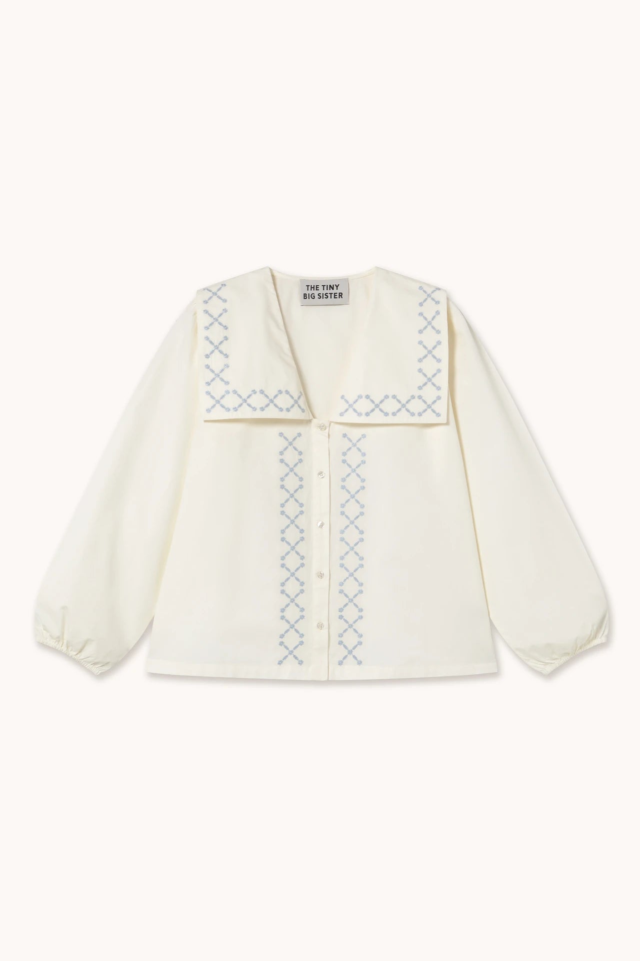 The Flower Embroidered Shirt - Ivory by Tiny Big Sister is a white cotton poplin blouse featuring a wide sailor collar and light blue cross-stitch embroidery, ideal for fans of sustainable fashion.