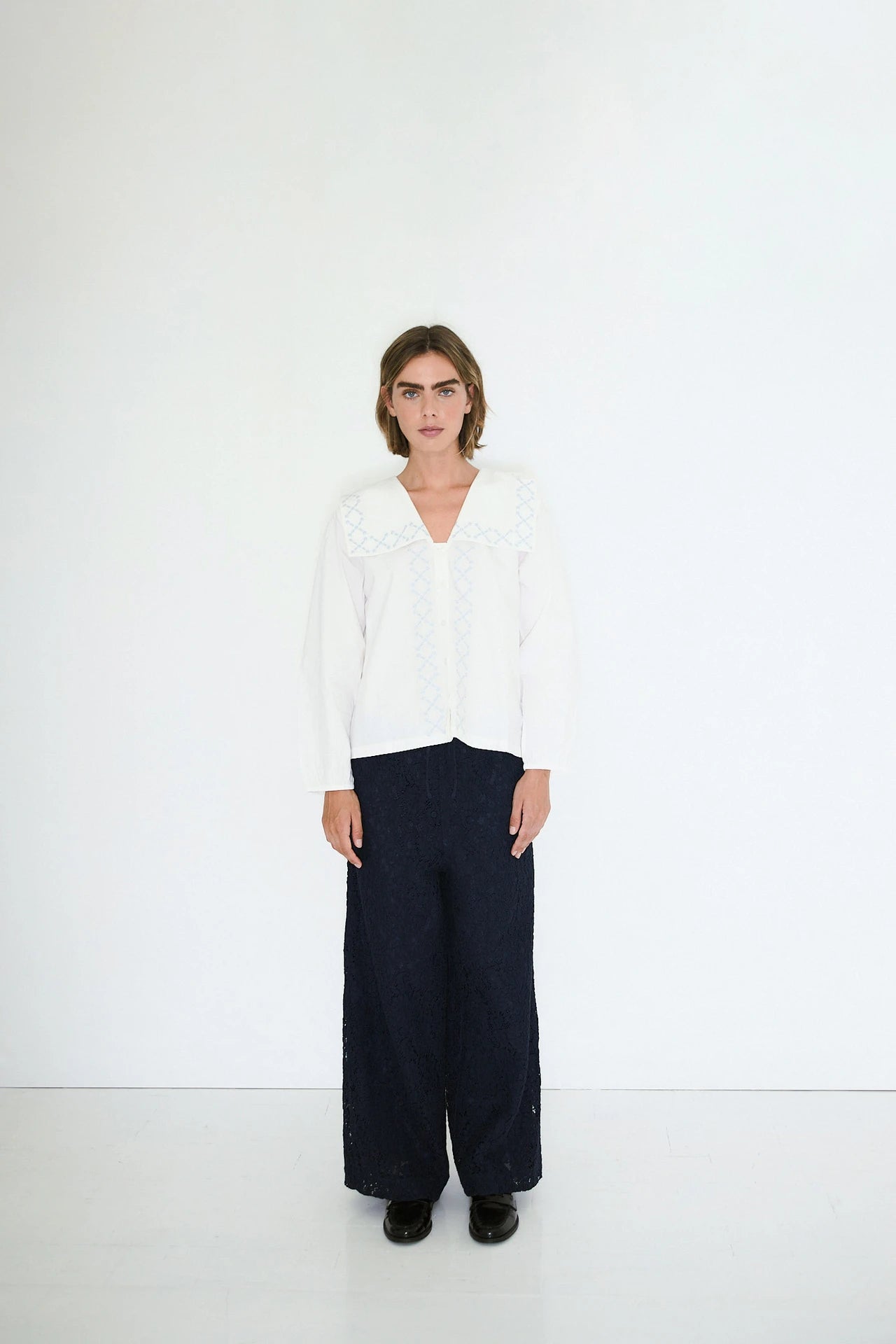 A person stands against a plain white background, wearing the Tiny Big Sister Flower Embroidered Shirt in ivory with black wide-leg pants and black shoes.