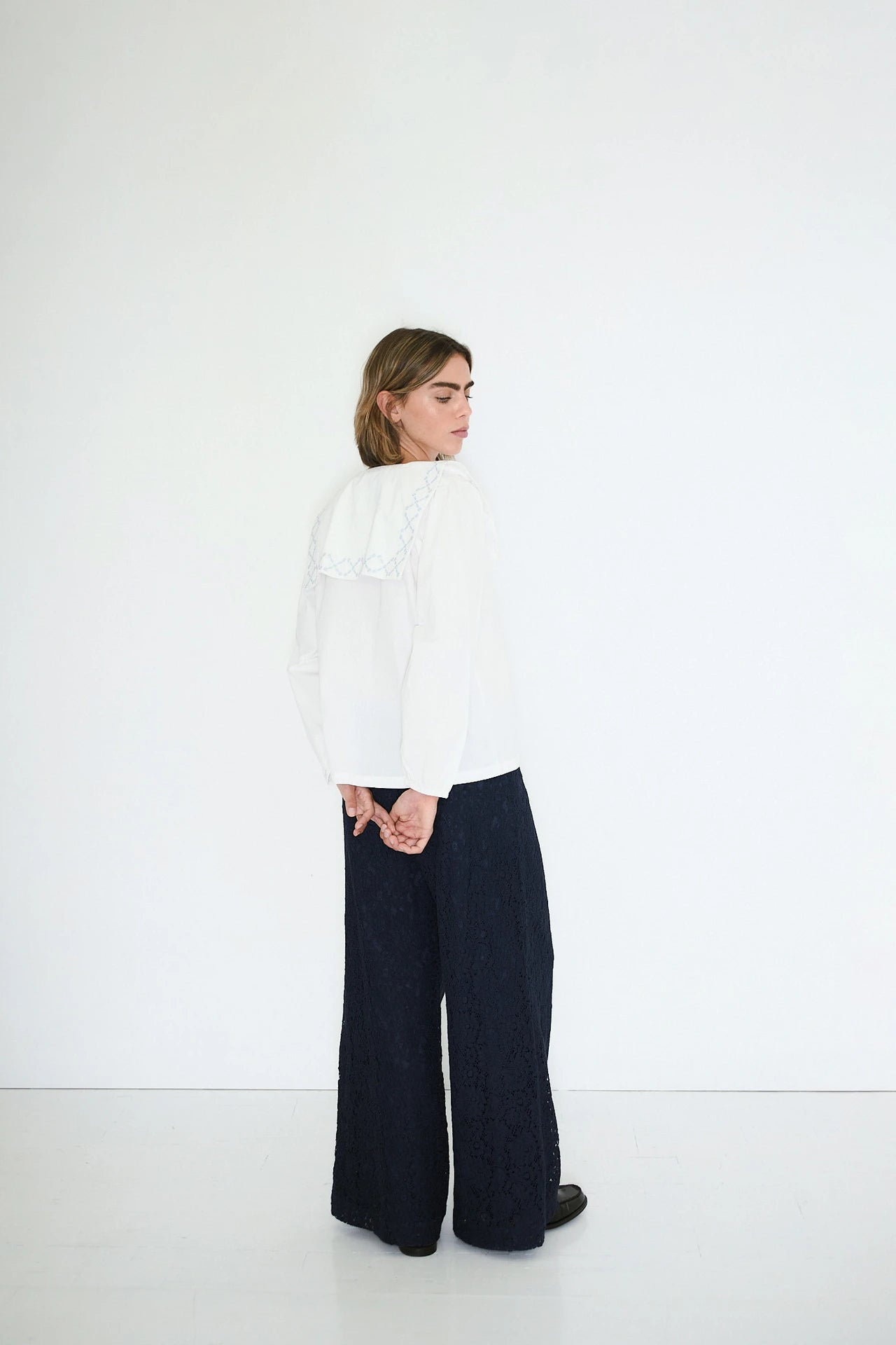 A person stands sideways against a white wall, hands behind their back, wearing Tiny Big Sister’s Flower Embroidered Shirt in ivory and wide-leg dark pants—a refined nod to sustainable fashion.