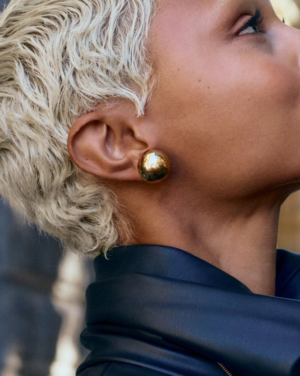 Close-up of a person with short blonde hair wearing EDBLAD's Bead Studs, crafted in shiny 14K gold plating, paired with a dark jacket—their lightweight design enhancing the elegance.