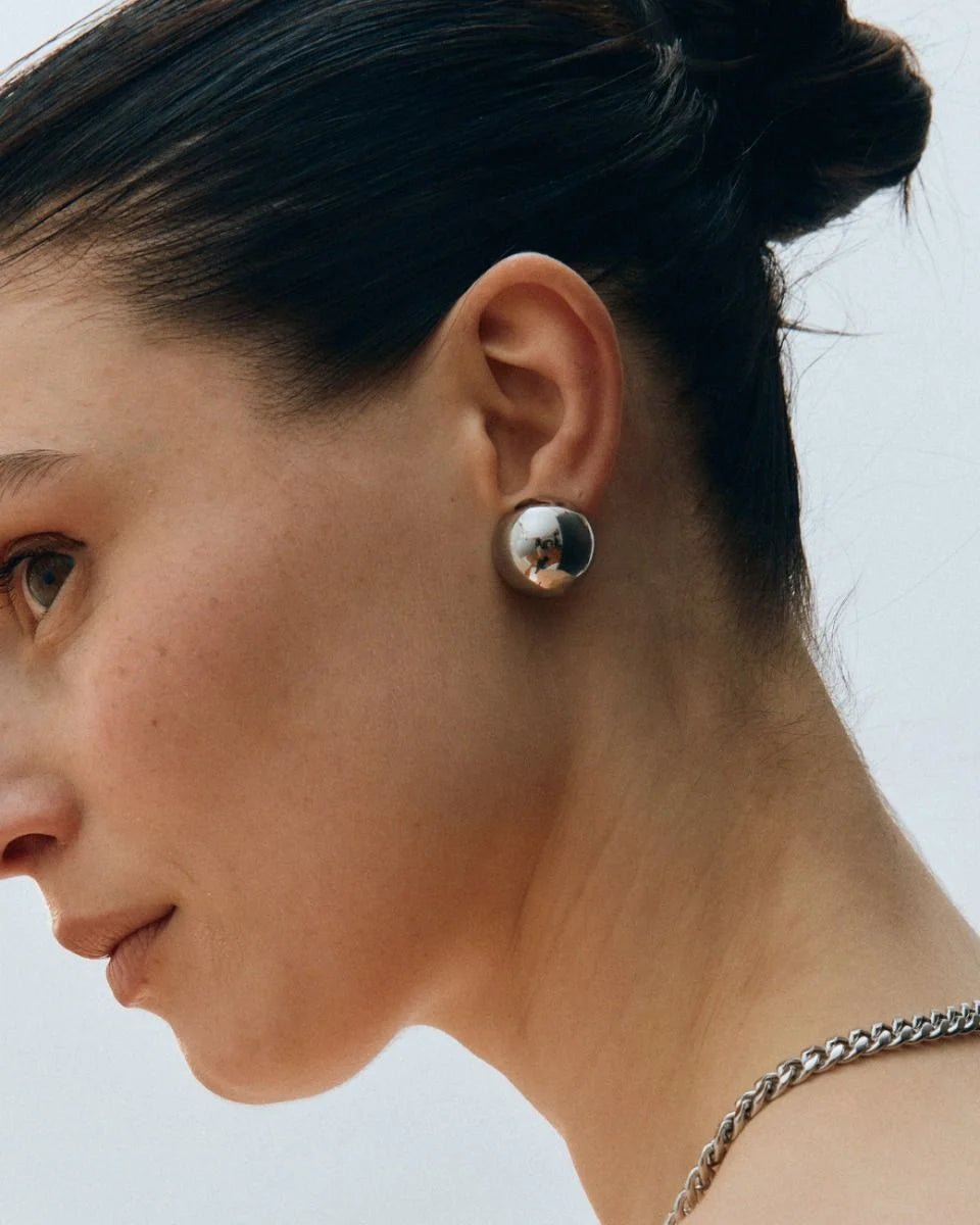 A person with short, dark hair wears the Bead Studs, a round, metallic earring from EDBLAD featuring a lightweight design, along with a silver necklace. They are facing sideways against a plain background.