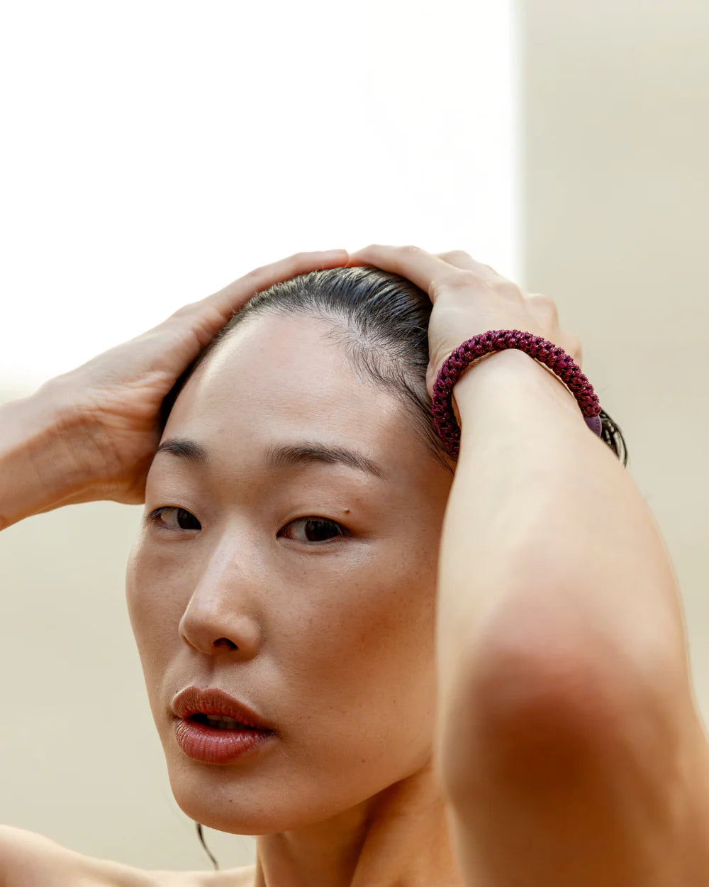 Someone with wet hair pulled back by a KKNEKKI Hair Tie - Original is gently holding their head, sporting a maroon beaded bracelet and gazing just past the camera.