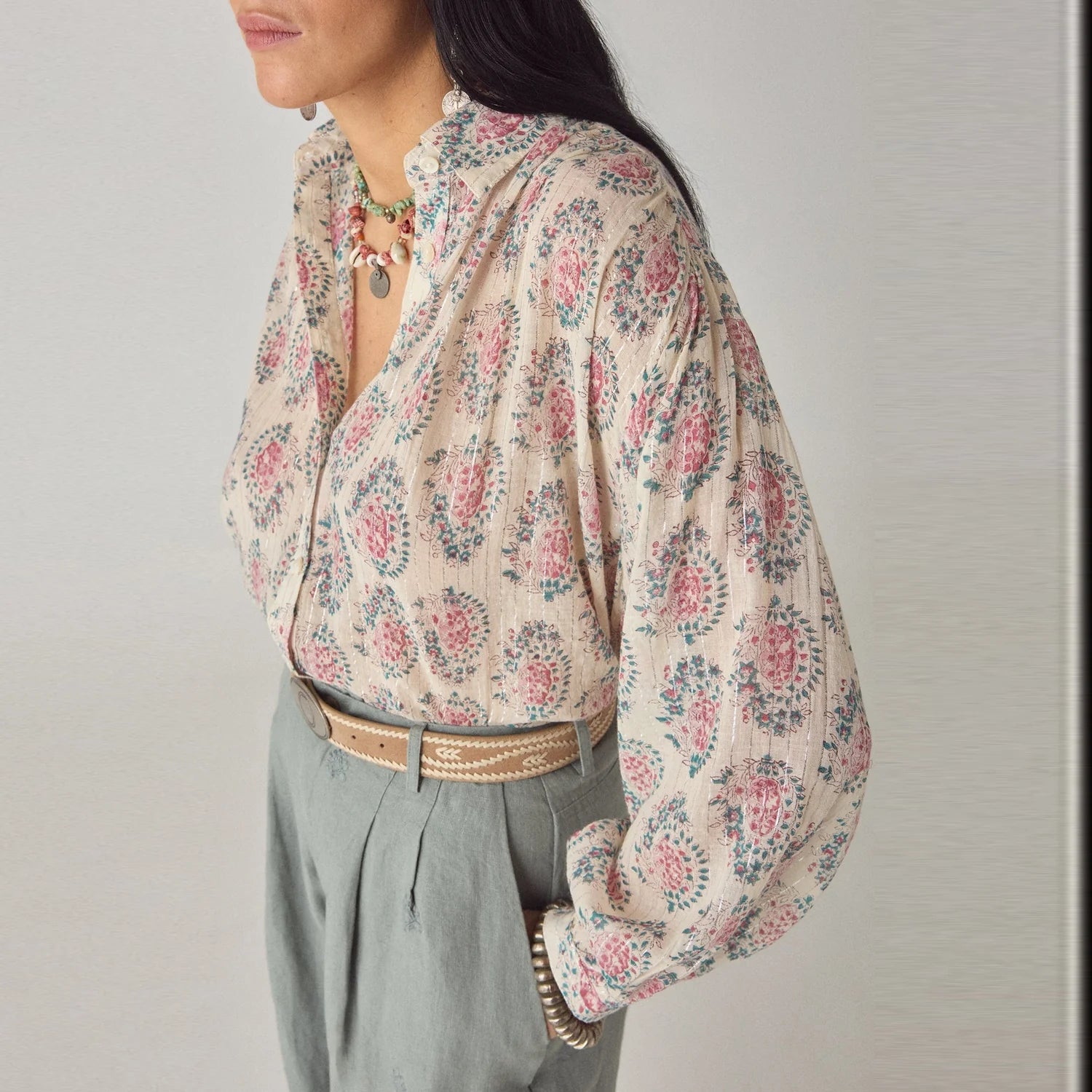 A woman wearing the Mary Blouse - Ivory by Maison Hotel, paired with high-waisted gray trousers, a braided belt, and a beaded necklace, stands against a plain background.