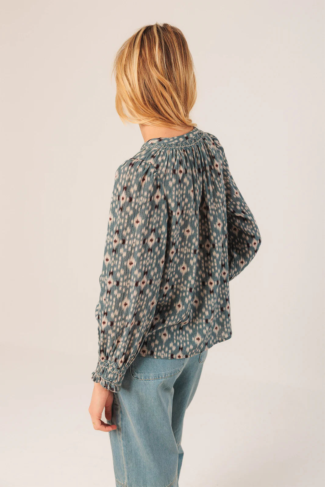 A person with blonde hair, wearing the Indi & Cold Smock Detail Shirt in Vintage Blue and blue jeans, stands facing away from the camera against a plain background.