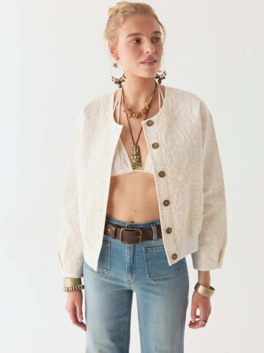 A woman embodies timeless elegance in a Tesela Ivory Boticcelli Jacket from Maison Hotel, paired with a white top and jeans. Her look is enhanced by large earrings, necklaces, and bracelets against a plain background.