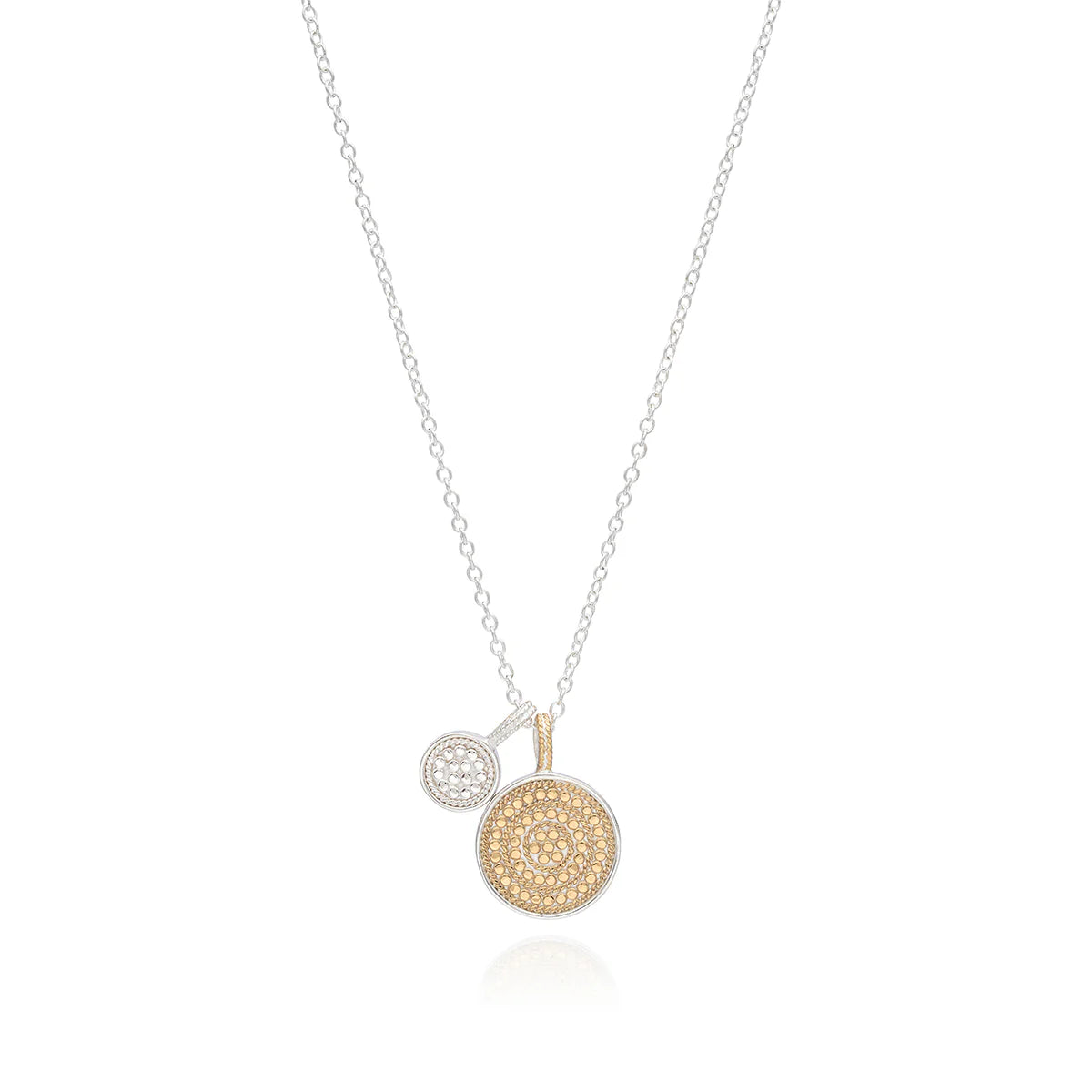 The Circle of Life Dual Disc Necklace by Anna Beck is a silver chain necklace featuring two circular pendants: a larger one with an 18k gold finish and a smaller one with a silver finish, both adorned with crystals. The necklace showcases a stunning two-tone design, highlighted by delicate honeycomb patterns on the pendants.