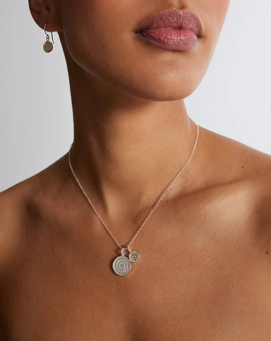 A person is adorned with the "Circle of Life Dual Disc Necklace" by Anna Beck, which showcases two circular pendants with an intricate honeycomb pattern, paired with matching earrings.