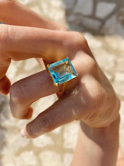 The SHYLA - Claudia Ring - Gold, adorned with a beautiful blue topaz stone, exudes a vintage look as it delicately rests in the palm of a woman's hand. (Brand Name: Shyla)