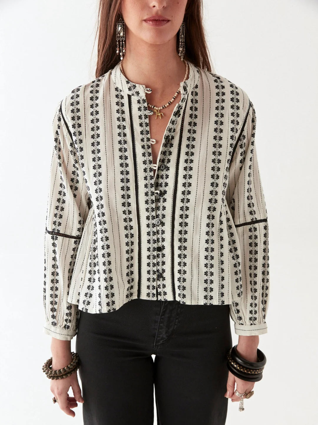 Dressed in a Maison Hotel Consuelito Blouse - Pearl Jam and black pants, a person stands against a plain backdrop, accessorized with bracelets and a necklace for extra flair.