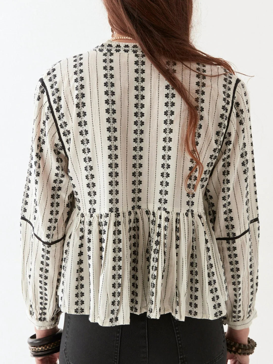 A person with long hair wears the Consuelito Blouse - Pearl Jam by Maison Hotel, showcasing a patterned, long-sleeve design with ruffled trim and black embroidery, viewed from the back.