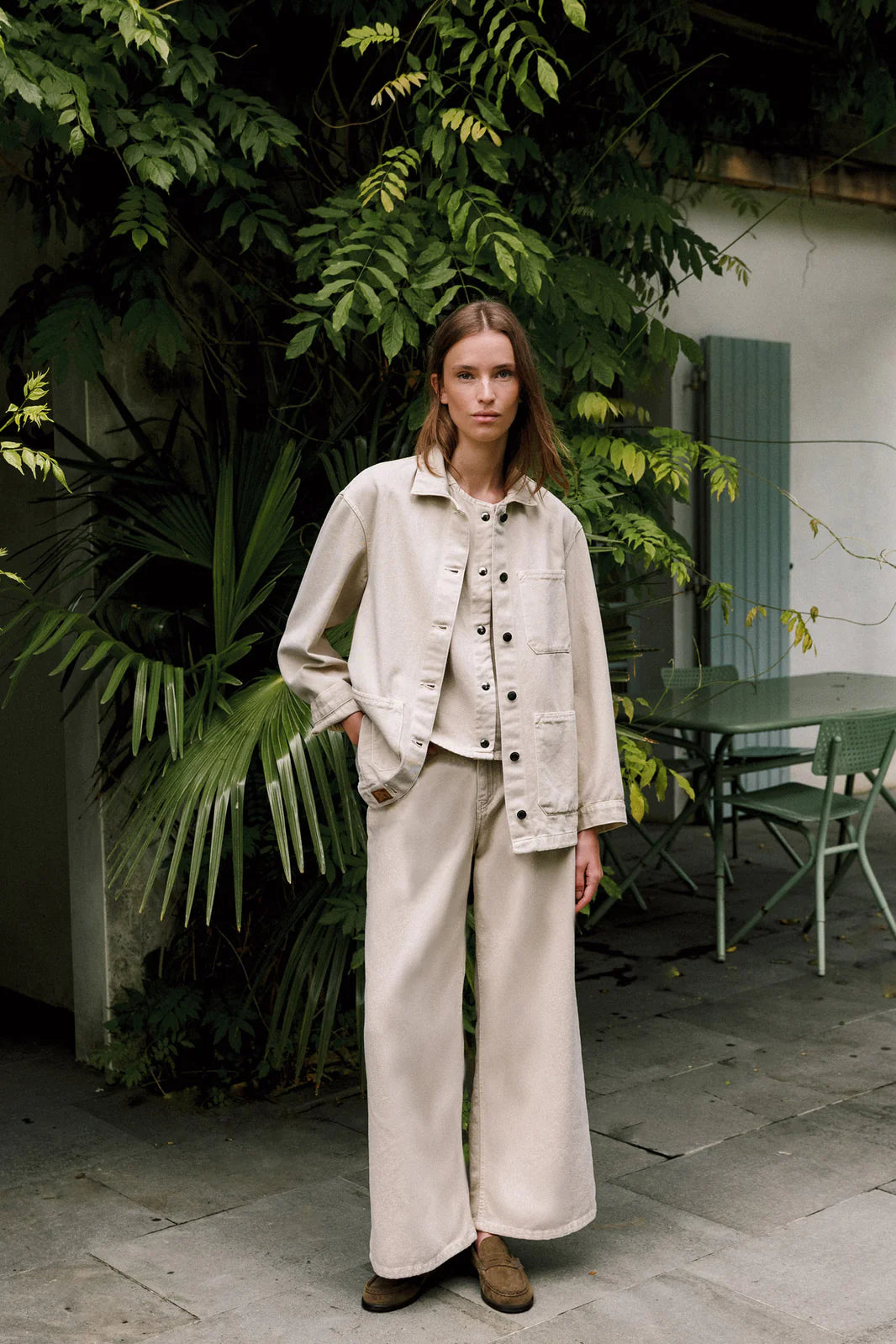 A woman stands outdoors by leafy plants, wearing Indi & Cold's Wide Twill Trousers in cream with a loose beige jacket and brown shoes. Green patio furniture is visible in the background.