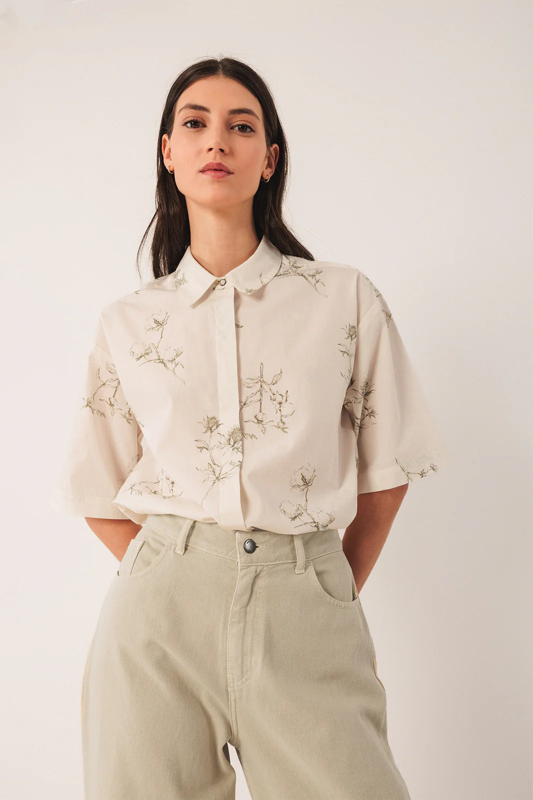 A woman stands against a plain background wearing Indi & Cold's Wide Twill Trousers in Cream, paired with a light, short-sleeve floral button-up shirt, her hands tucked casually in the pockets.