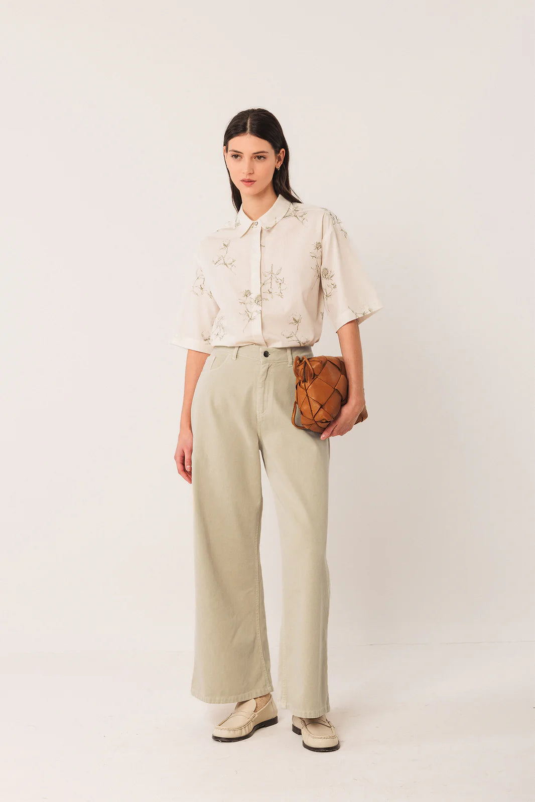 A woman stands against a white background wearing a floral blouse, cream Wide Twill Trousers by Indi & Cold, cream shoes, and holding a brown quilted clutch.