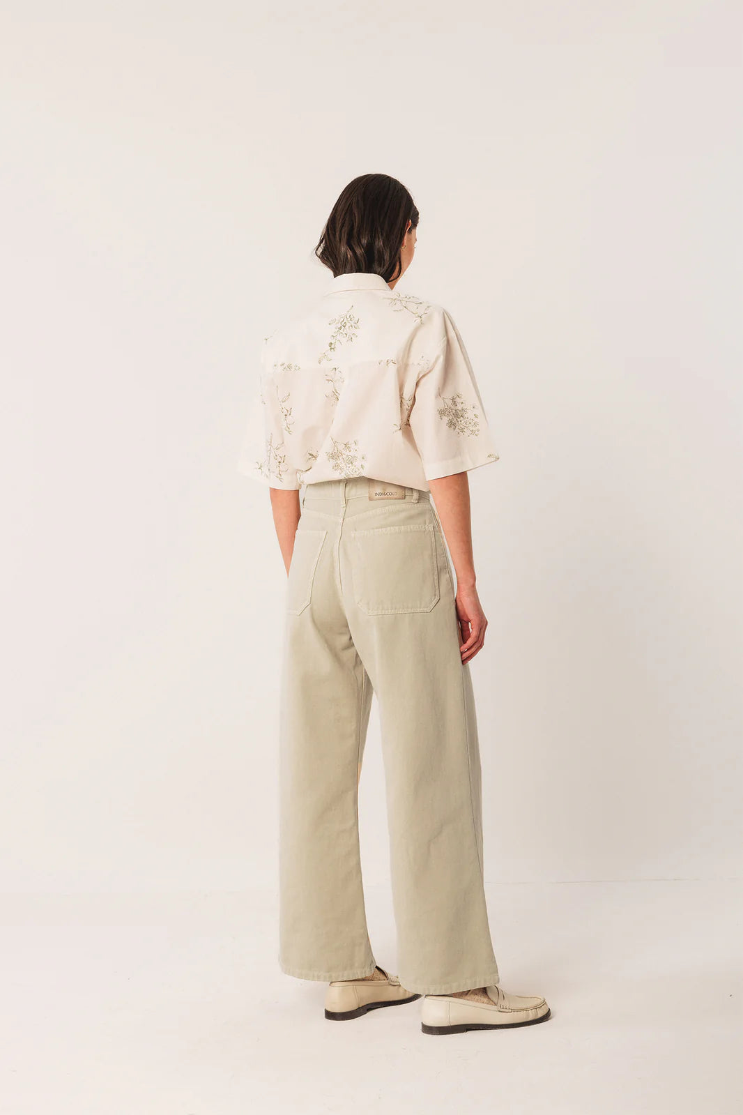A person stands in a neutral studio, facing away, styled in a short-sleeved patterned shirt with Indi & Cold’s Wide Twill Trousers in Cream and light-colored shoes.