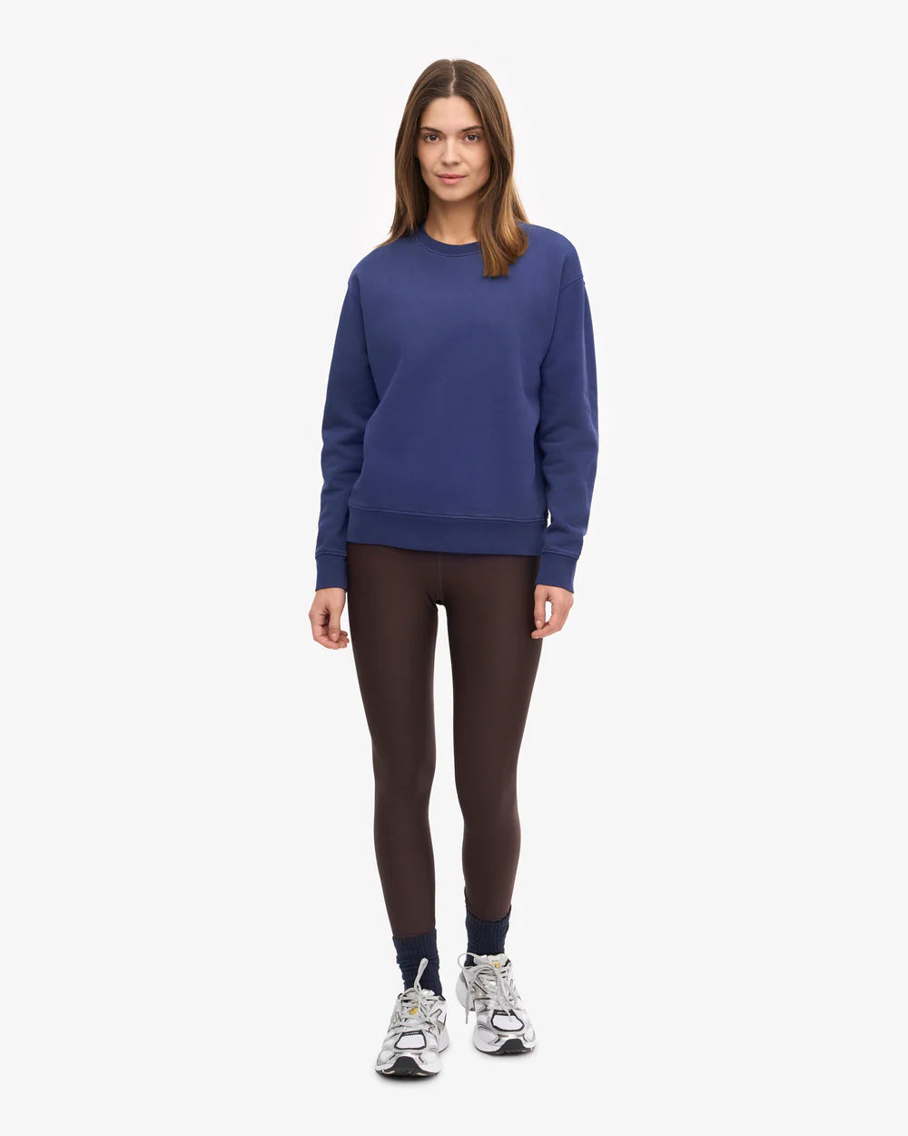 A woman faces forward wearing the Colorful Standard Classic Organic Crew in blue, dark brown leggings, navy socks, and gray sneakers against a plain white background.