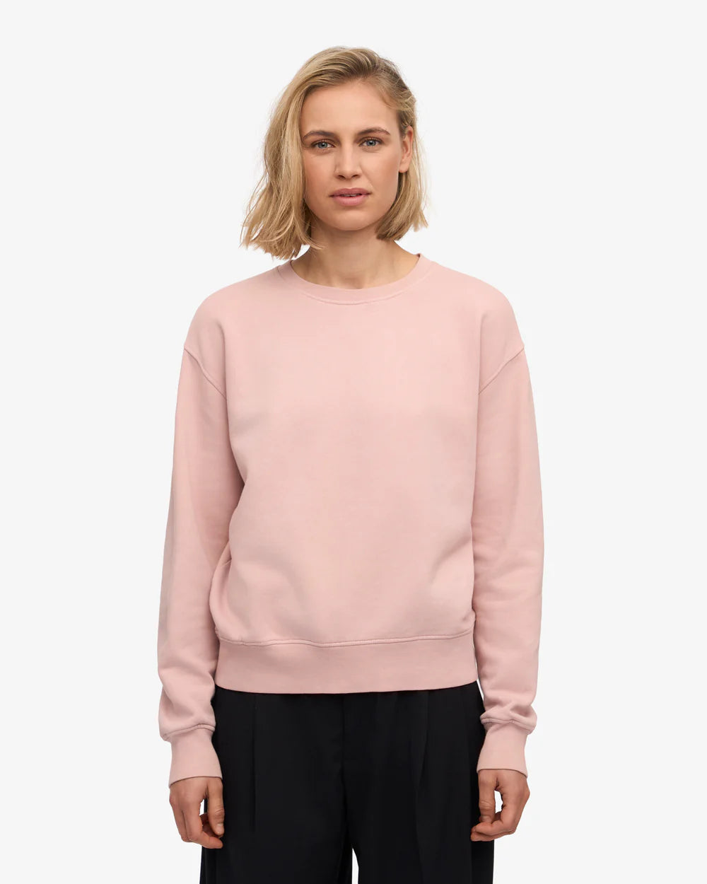 A person with short blonde hair wears the Colorful Standard Classic Organic Crew in light pink with black pants, standing against a plain white background.
