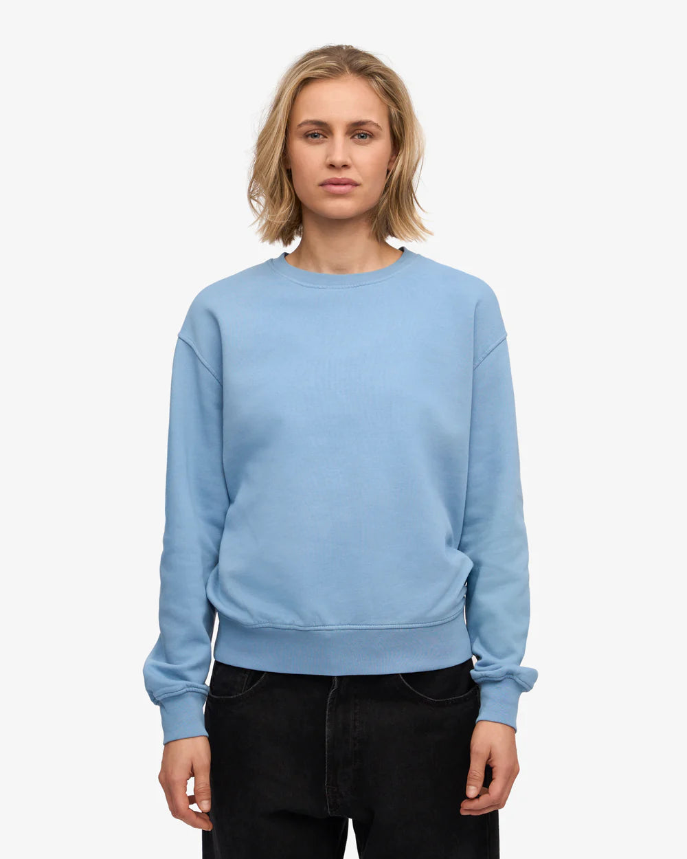 A person with blonde hair wearing the Colorful Standard Classic Organic Crew in light blue and black pants stands against a plain white background.