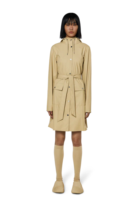 A person stands facing forward against a plain white background, wearing the Rains Curve W Jacket W3—a beige women's raincoat with a belt—paired with beige knee-high socks and beige shoes.