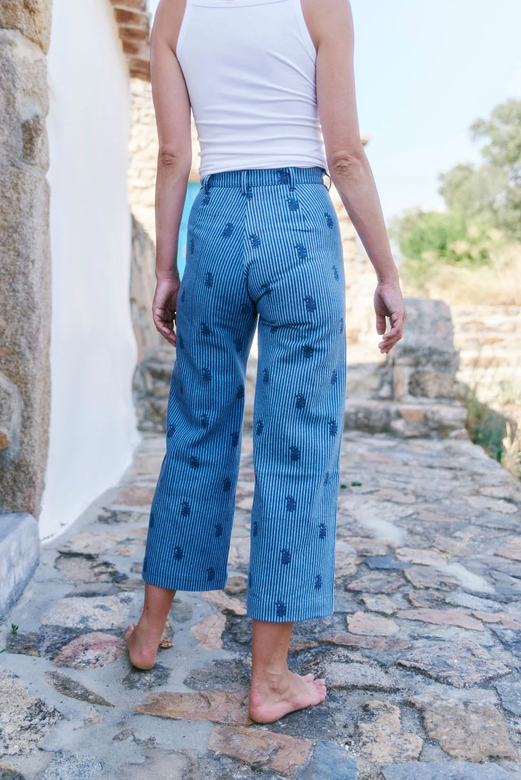 A person wearing SZ Blockprints' Disco Pant - Denim Paisley, barefoot in a white tank top, stands on a stone path by a building with a stone wall.