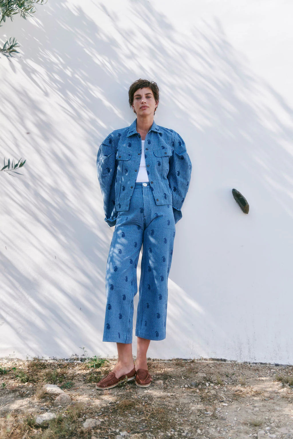 A person wearing the Disco Pant - Denim Paisley by SZ Blockprints stands against a white wall with tree shadows.