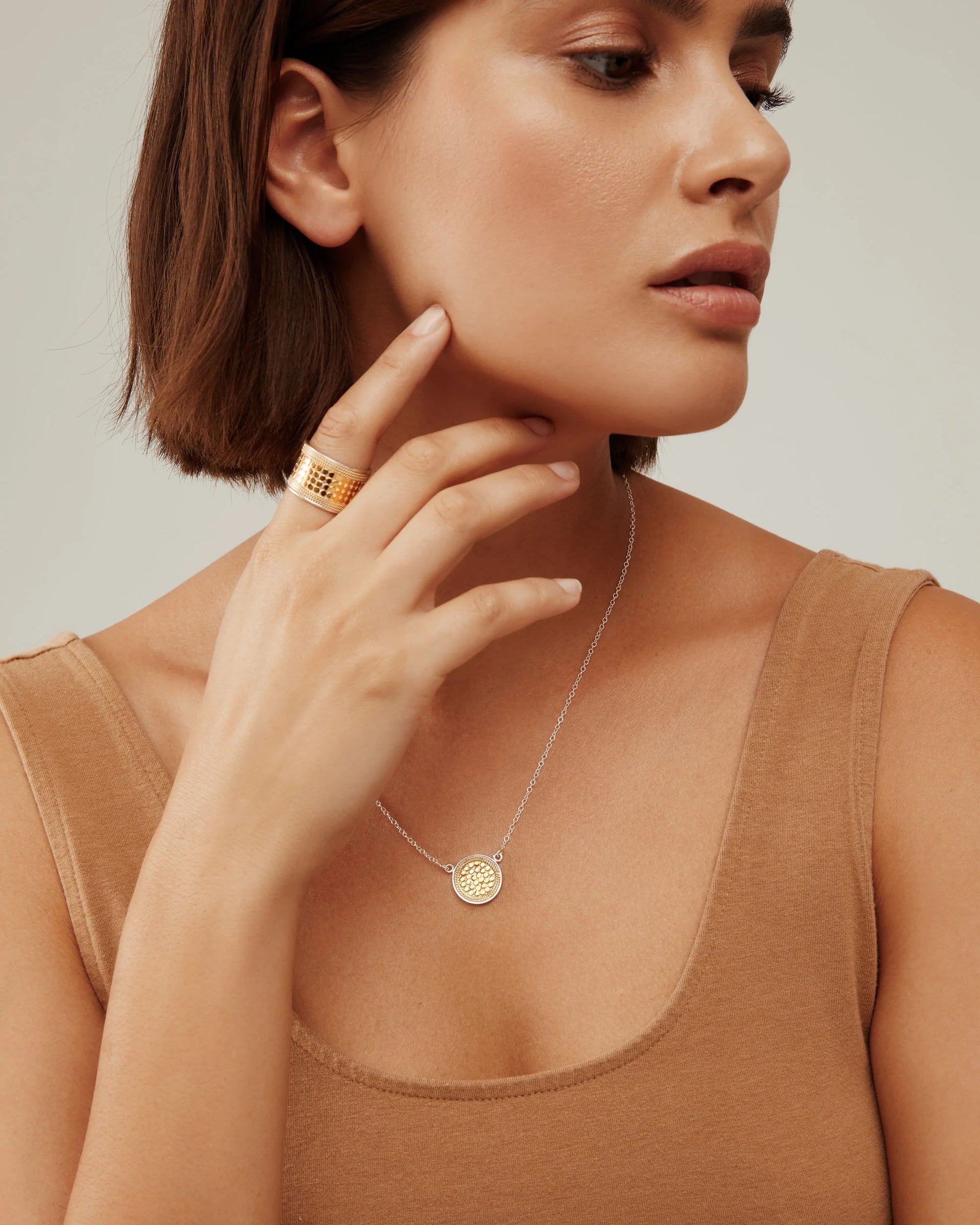 A woman with short brown hair wearing a beige sleeveless top, a gold ring, and the Classic Disc Necklace in gold by Anna Beck touches her face thoughtfully.