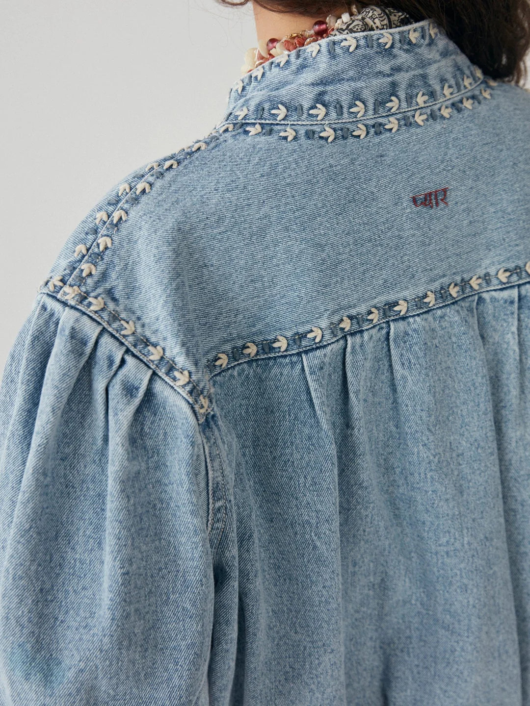 Model wears denim jacket with round collar and embroidered seams and cuffs