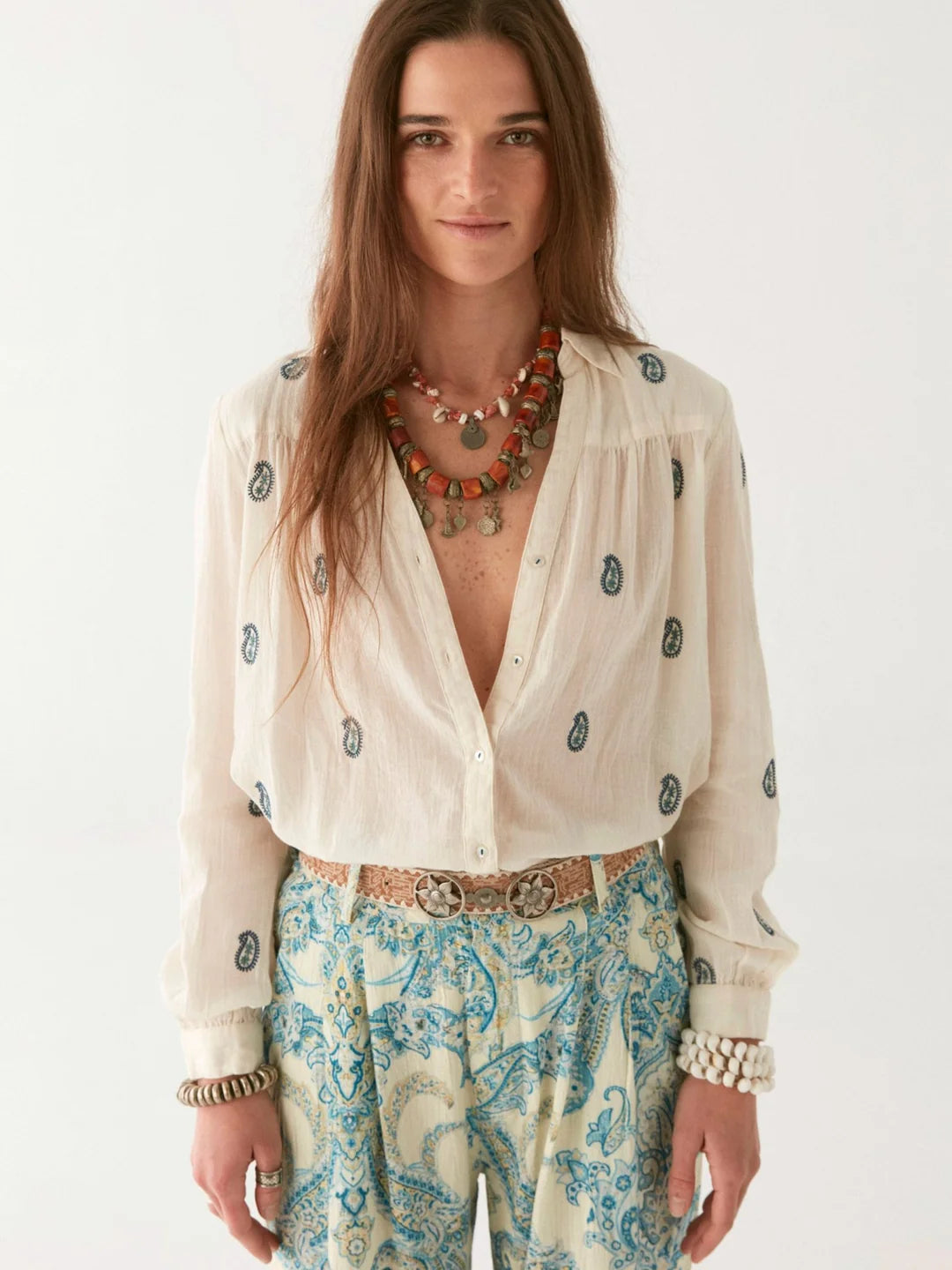 A woman with long hair, wearing a Maison Hotel ivory Dylan Blouse with paisley embroidery, paired with beaded necklaces and paisley pants, stands against a plain background.