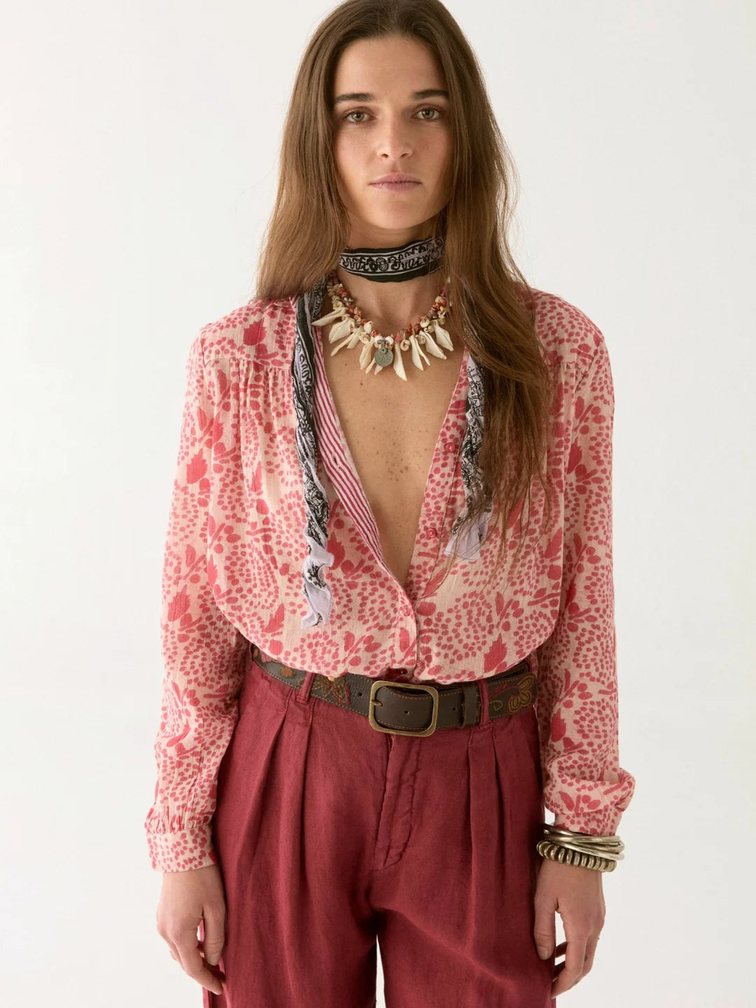 A woman with long brown hair wears a Dylan Blouse by Maison Hotel, featuring a red floral pattern, maroon pants, and layered necklaces. She accessorizes with bracelets and a belt, standing confidently against a plain backdrop.