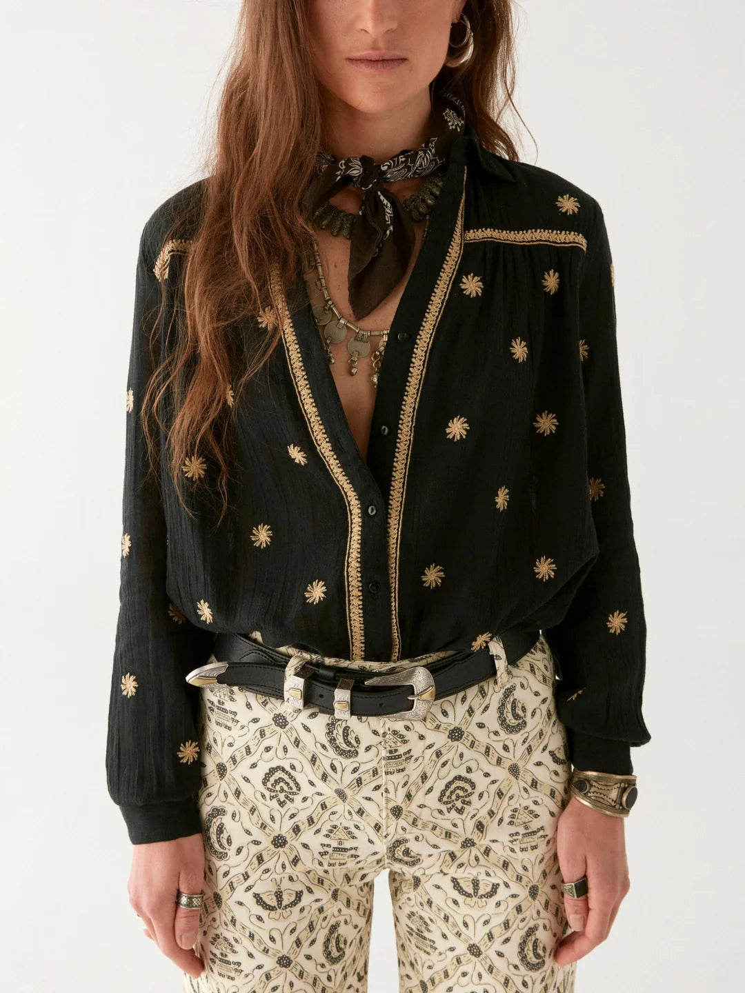 A person dons a black, starry-patterned Dylan Blouse from Maison Hotel, paired with white patterned pants and a scarf. The look is accessorized with a necklace, rings, and bracelets for a bohemian vibe.