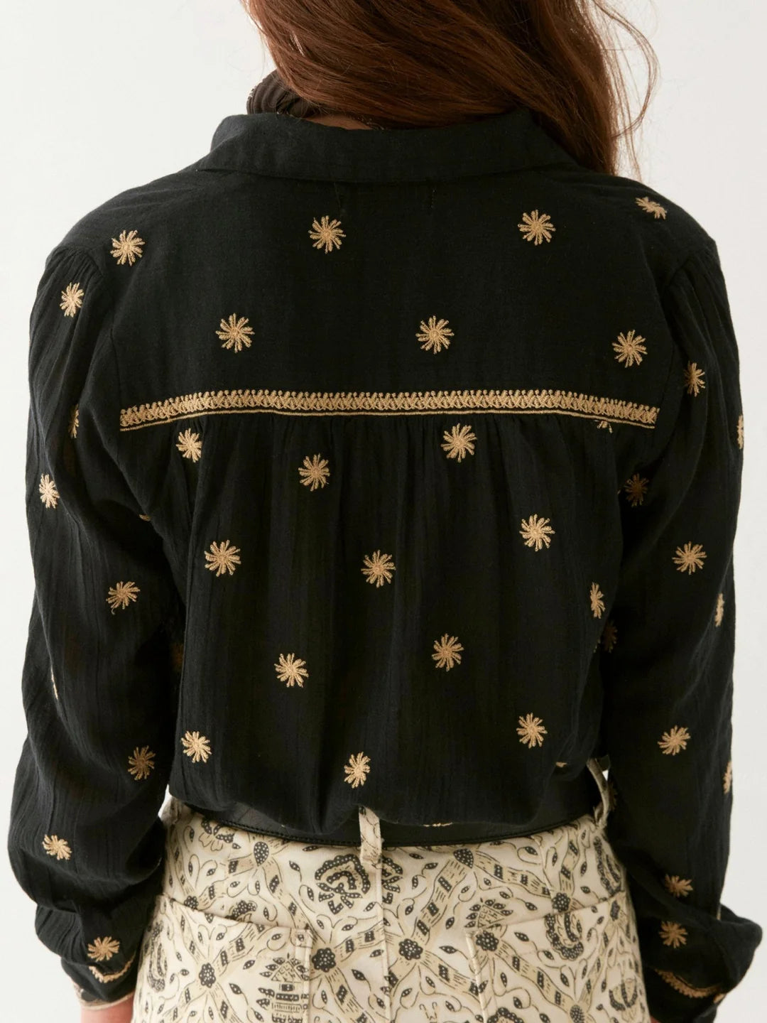 A person with long hair wears the Maison Hotel Dylan Blouse - Starry Night, showcasing gold embroidered suns, paired with light pants with a black motif. The view from the back captures a bohemian essence effortlessly.