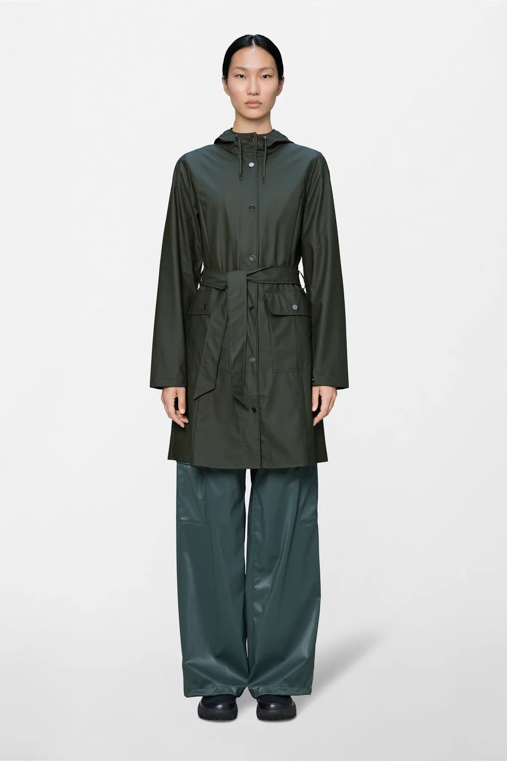 A person is wearing the Rains Curve W Jacket W3, a dark green women’s raincoat with a hood and waist belt, paired with wide-legged waterproof pants and black shoes, standing against a plain light background.
