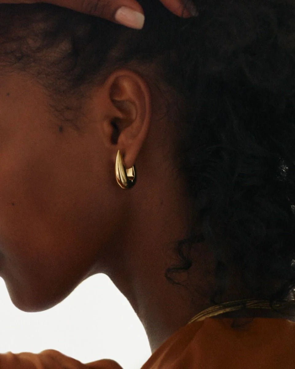 Close-up of a person wearing EDBLAD's Crest Hoops L, 14K gold-plated crescent-shaped earrings, with a hand resting on their head and curly hair partially visible.