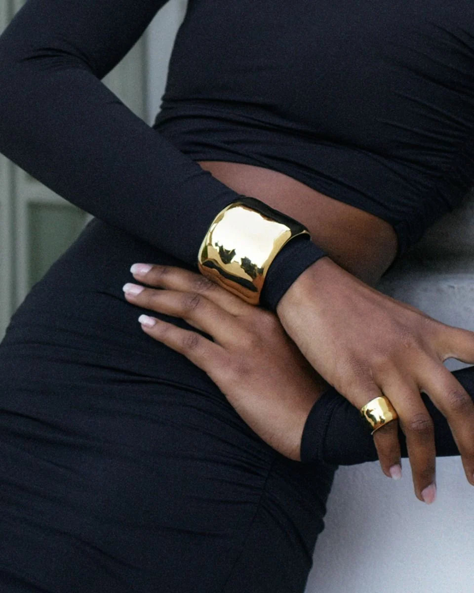 A person wearing a long-sleeved black dress poses with statement jewellery, including EDBLAD's Flowy Bangle and ring. The eye-catching design features 14K gold plating, with their arms crossed over their waist.