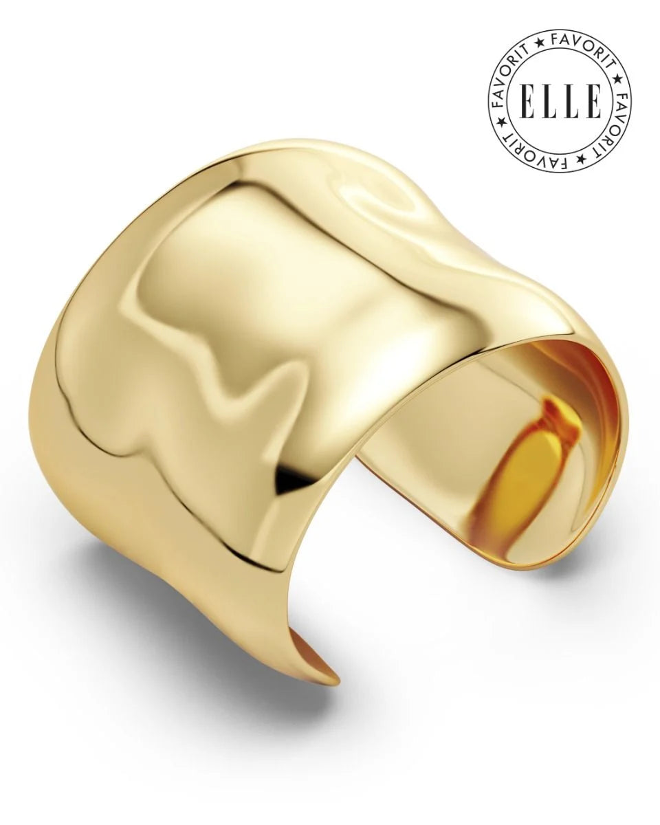 A wide, shiny gold Flowy Bangle by EDBLAD with 14K gold plating and a smooth, reflective surface is shown. The 'ELLE Favorite' stamp is in the top right corner, highlighting its eye-catching design.