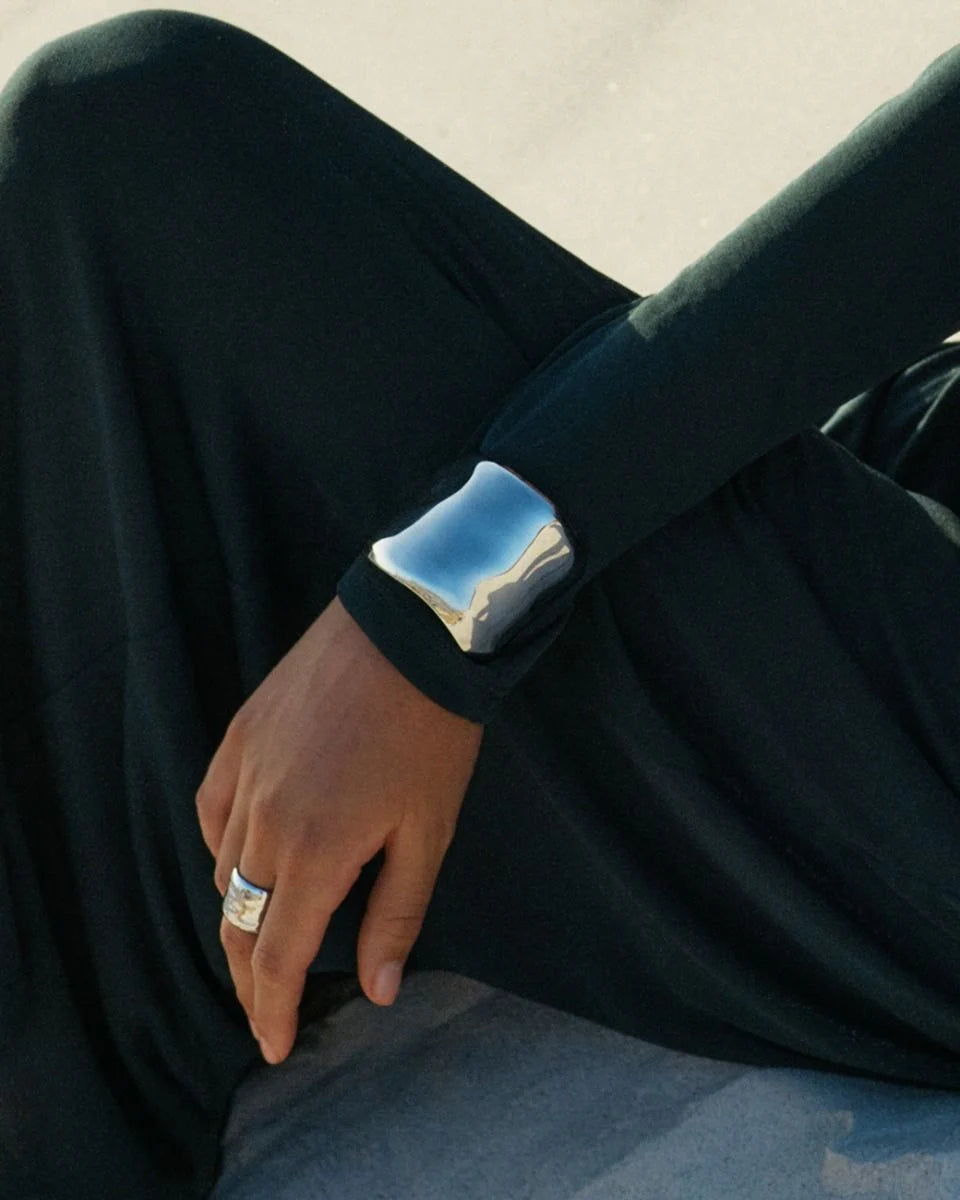 Person wearing the Flowy Bangle and a silver ring on their left hand, resting on dark fabric, showcasing EDBLAD statement jewellery with an eye-catching design.