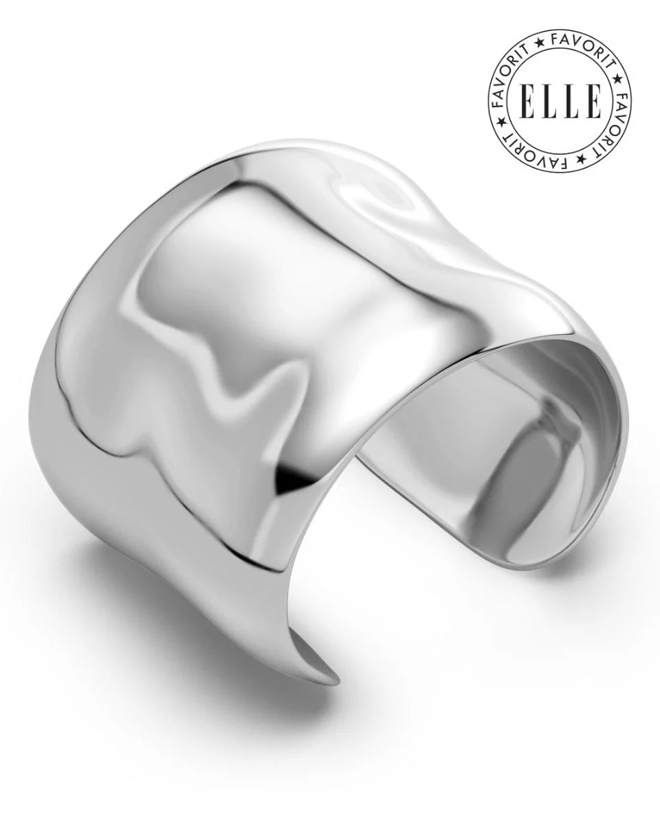 The EDBLAD Flowy Bangle is a wide, shiny silver cuff bracelet with smooth, curved edges and an "Elle Favorite" logo in the upper right corner. This eye-catching design exudes elegance and sophistication.