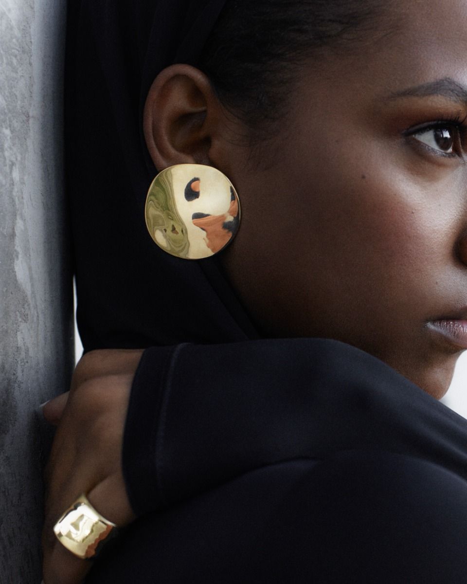 A close-up of a person wearing EDBLAD's Flowy Studs Maxi, large round gold earrings and a gold ring, both showcasing bold metal jewelry with 14K gold plating, along with a portion of their face and black clothing visible.