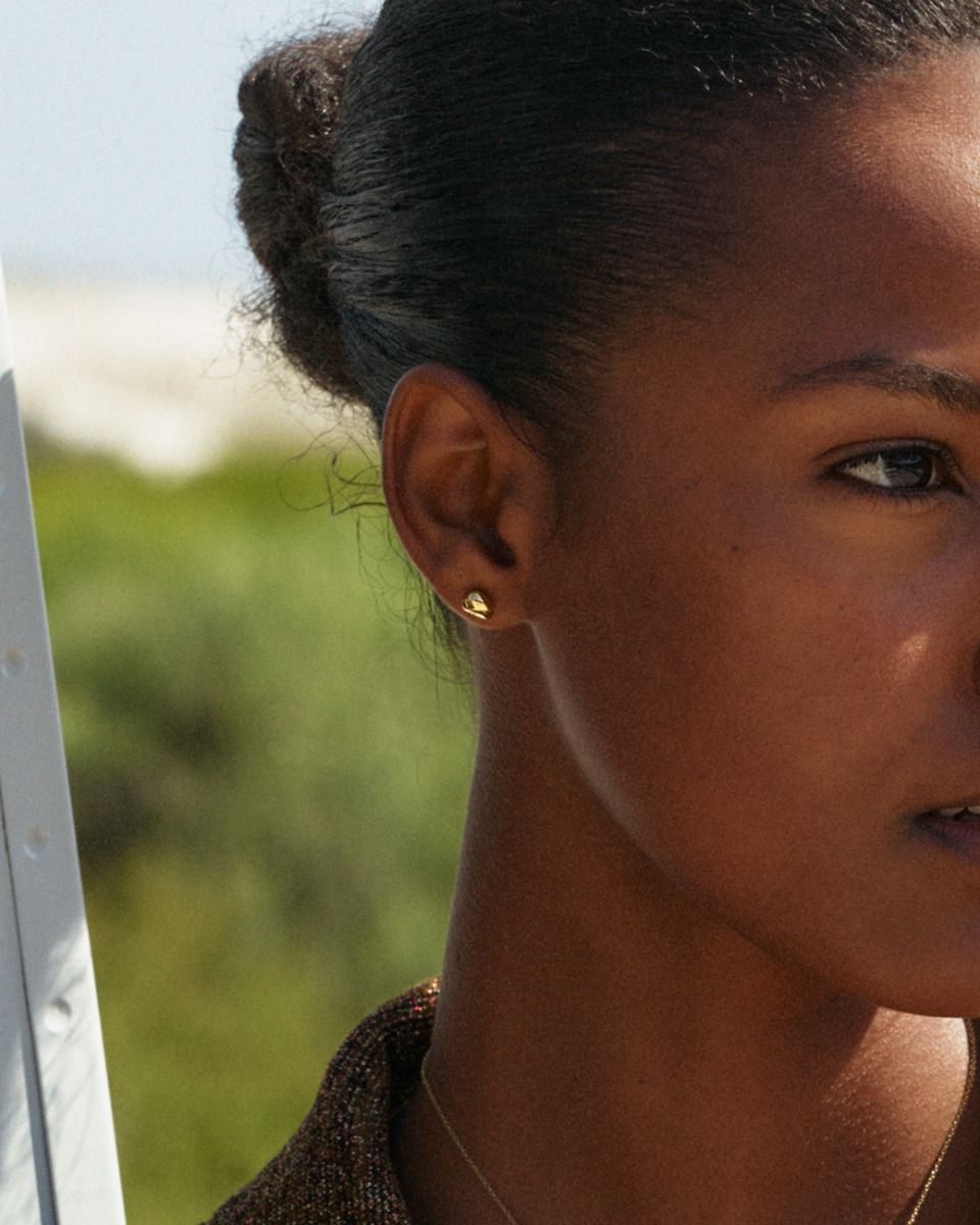 Partial view of a person with a braid wearing the Lulu studs - Gold by EDBLAD, standing outdoors with greenery in the background. Perfect for everyday wear, the 14K gold plating adds an elegant touch to this modern sculptural drop design.