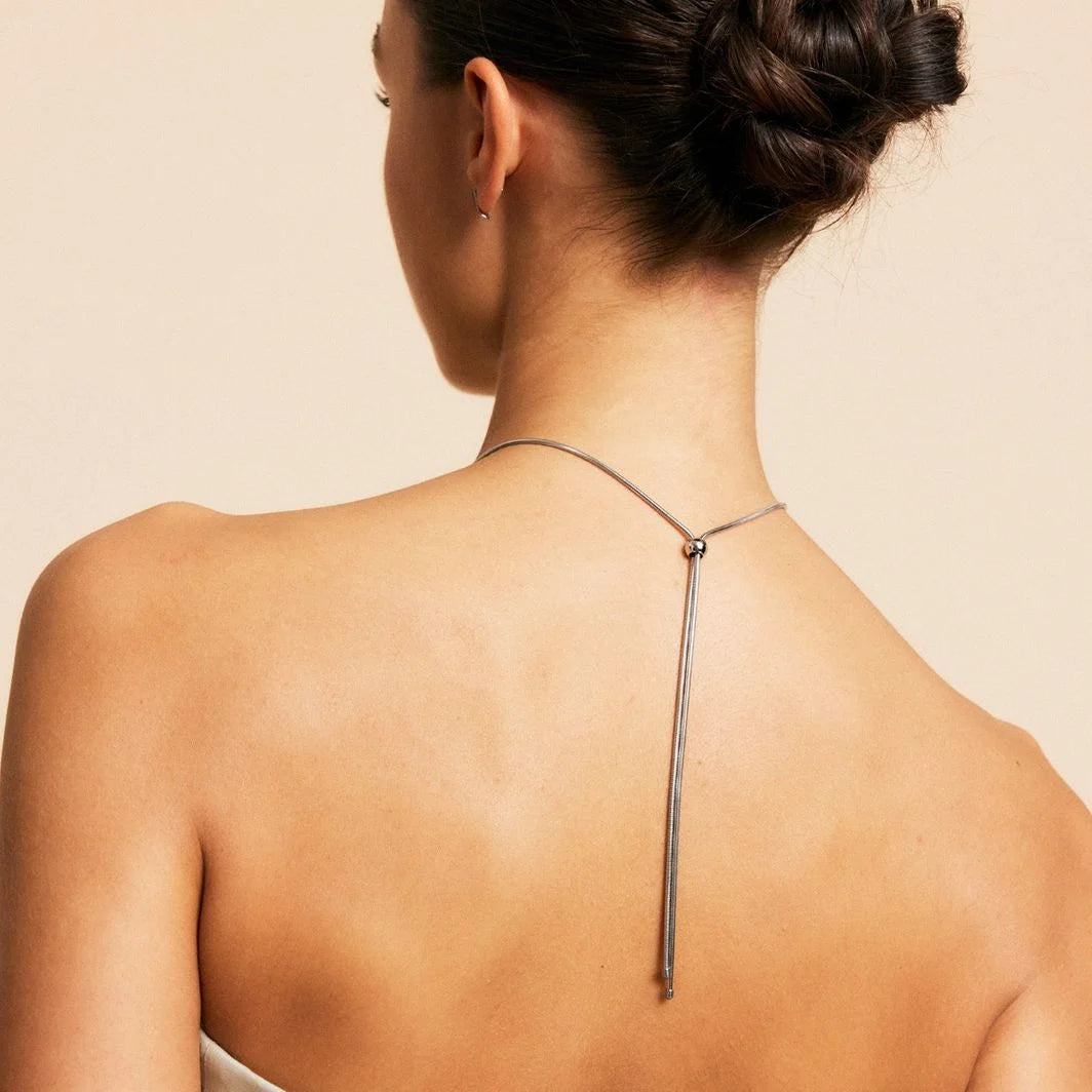 A woman with her hair styled in an updo is wearing a backless dress and the Melrose Necklace L from EDBLAD, featuring 14K gold plating on a long chain that elegantly hangs down her back.