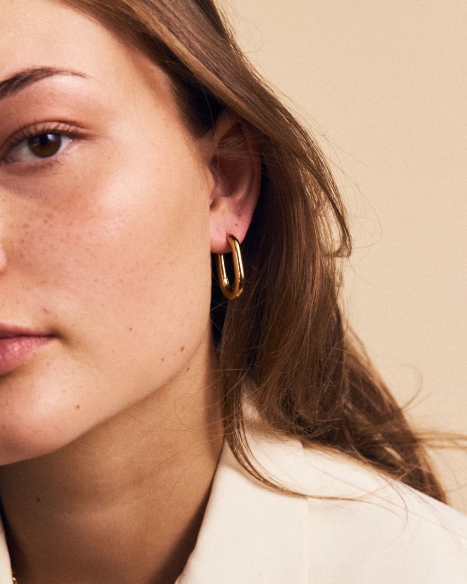 Close-up of a person with long brown hair, wearing a single EDBLAD Modernist Hoops L earring in 14K gold plating, against a beige background.